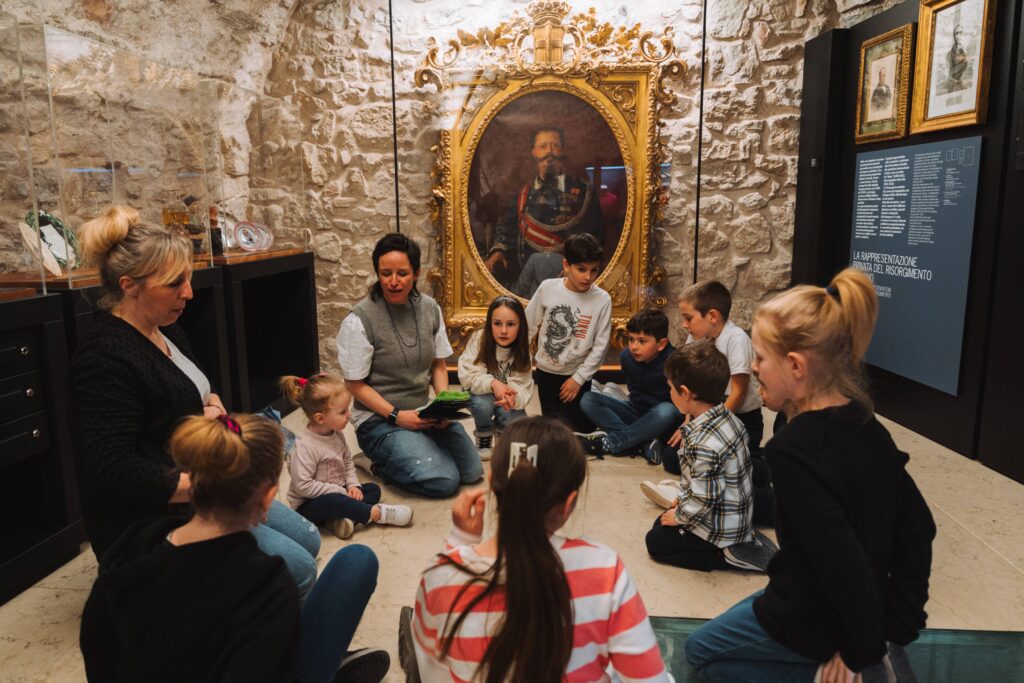 Bambini e genitori seduti a terra ascoltano un'educatrice in una sala del museo