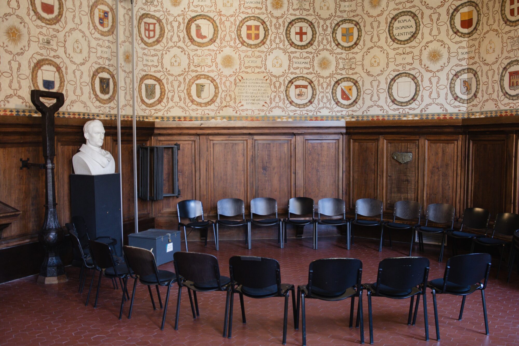 Sala storica con pareti decorate a stemmi, un busto e sedie disposte in cerchio