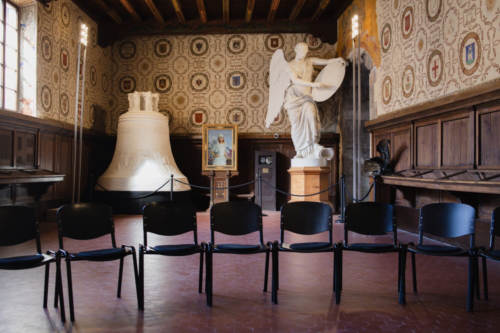 Sala con pareti decorate a stemmi, una grande campana e una statua di angelo